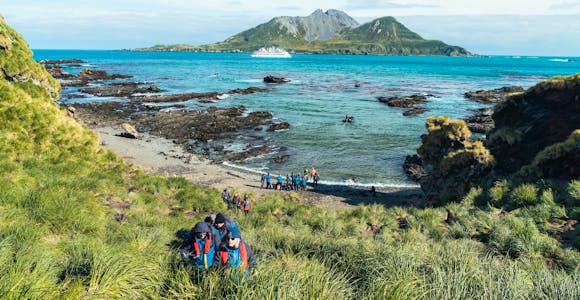 A group of tourists climb a grassy hill up from a beach at Cooper Bay in South Georgia. A zodiac drives to a cruise ship is anchored in the bay. 