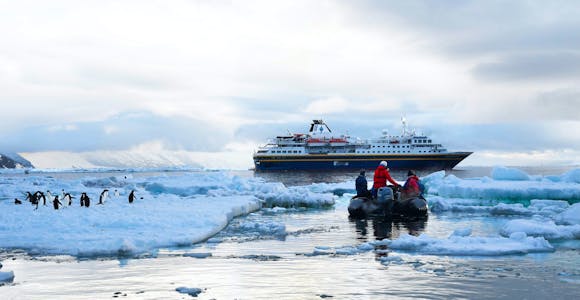 A zodiac boat carrying tourists cruises past an ice floe with Adelie penguins in the Ross Sea. The ship MS Adventurer is on the horizon