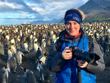 A female tourist in a blue jacket wearing a Swoop Adventures buff and carrying a camera stands smiling in front of thousands on king penguins of South Georgia island