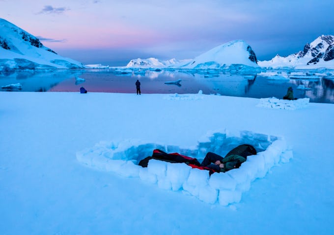 Campers camp in a snowhole in Antarctica while a pink-tinged sun sets in the background