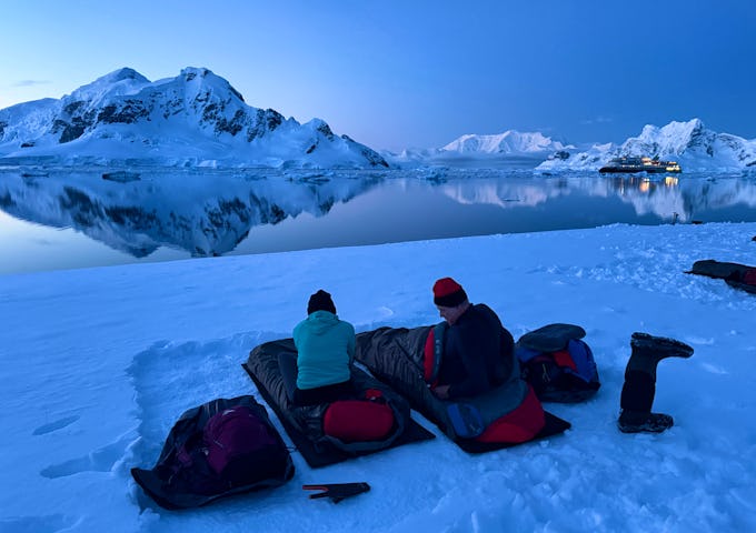 Two people sit on sleeping bags in a dusky Antarctic bay ahead of a night camping