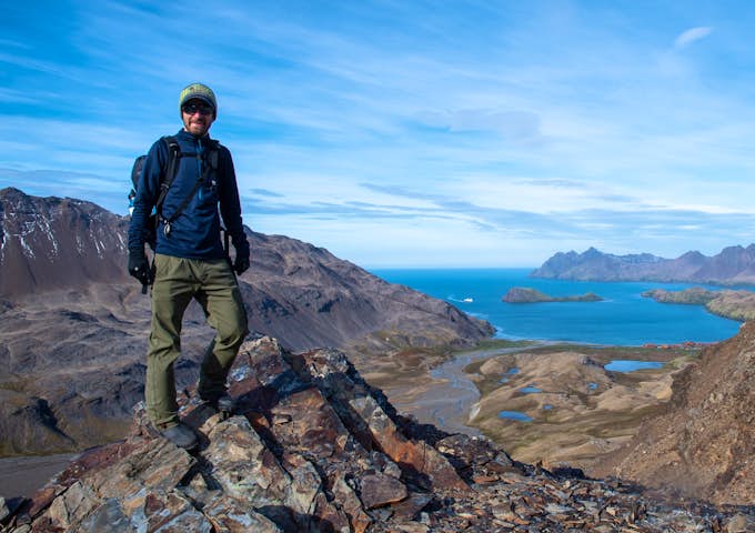 A man stands on a mountain ridge on South Georgia Island. Behind him is a distant blue bay with Stromness whaling station, at the end of the Shackleton hike